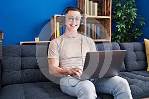 Young hispanic man using laptop and headphones sitting on sofa at home