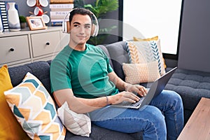 Young hispanic man using laptop and headphones sitting on sofa at home