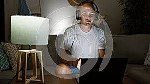 Young hispanic man using laptop and headphones sitting on sofa at home