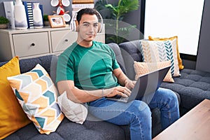 Young hispanic man using laptop and headphones sitting on sofa at home