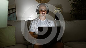 Young hispanic man using laptop and headphones sitting on sofa at home