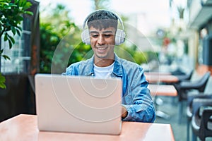 Young hispanic man using laptop and headphones sitting on table at coffee shop terrace