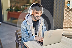 Young hispanic man using laptop and headphones sitting on table at coffee shop terrace