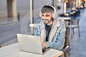 Young hispanic man using laptop and headphones sitting on table at coffee shop terrace