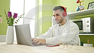 Young hispanic man using laptop and headphones sitting on table at dinning room