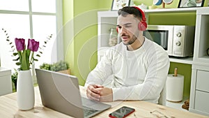 Young hispanic man using laptop and headphones sitting on table at dinning room