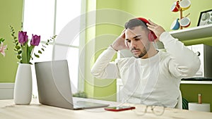 Young hispanic man using laptop and headphones sitting on table at dinning room