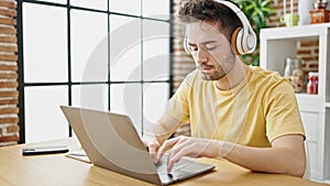 Young hispanic man using laptop and headphones sitting on table at dinning room