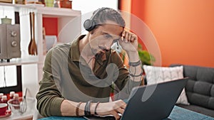 Young hispanic man using laptop and headphones tired at dinning room