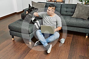 Young hispanic man using laptop sitting on the floor with dog at home