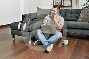 Young hispanic man using laptop and talking on the smartphone sitting on the floor with dog at home