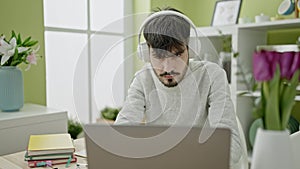 Young hispanic man using laptop wearing headphones at dinning room