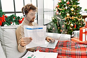 Young hispanic man using laptop working sitting by christmas tree at home