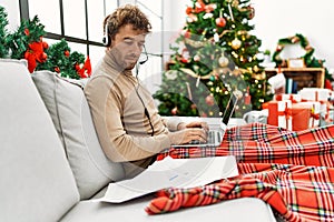 Young hispanic man using laptop working sitting by christmas tree at home