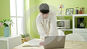 Young hispanic man using laptop writing on notebook at dinning room