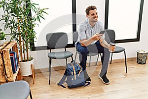 Young hispanic man using smartphone and drinking coffee sitting on chair at waiting room