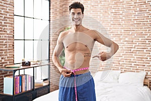 Young hispanic man using tape measure measuring waist smiling happy pointing with hand and finger