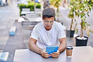 Young hispanic man using touchpad drinking coffee at coffee shop terrace
