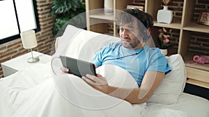 Young hispanic man using touchpad and headphones lying on bed at bedroom