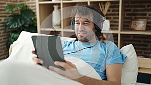 Young hispanic man using touchpad and headphones lying on bed at bedroom