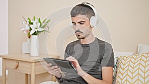 Young hispanic man using touchpad and headphones sitting on bed at bedroom