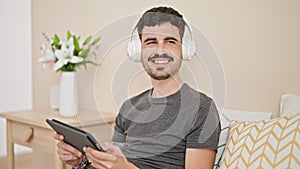 Young hispanic man using touchpad and headphones sitting on bed at bedroom