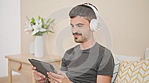 Young hispanic man using touchpad and headphones sitting on bed at bedroom