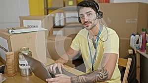 Young hispanic man volunteer using laptop and headphones smiling at charity center