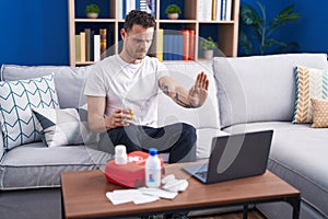 Young hispanic man watching video on laptop on how to use first aid kit with open hand doing stop sign with serious and confident