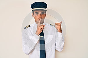 Young hispanic man wearing airplane pilot uniform asking to be quiet with finger on lips pointing with hand to the side