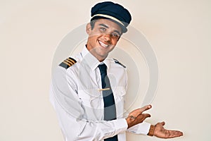 Young hispanic man wearing airplane pilot uniform inviting to enter smiling natural with open hand