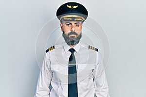 Young hispanic man wearing airplane pilot uniform relaxed with serious expression on face