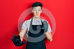 Young hispanic man wearing apron afraid and terrified with fear expression stop gesture with hands, shouting in shock