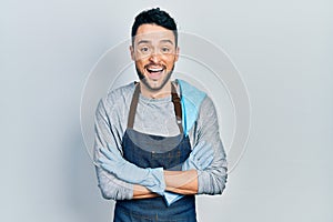 Young hispanic man wearing apron with arms crossed smiling and laughing hard out loud because funny crazy joke