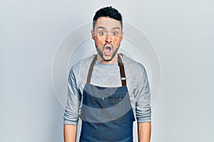 Young hispanic man wearing apron and gloves scared and amazed with open mouth for surprise, disbelief face