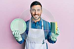Young hispanic man wearing apron holding scourer washing dishes smiling with a happy and cool smile on face