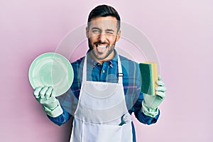Young hispanic man wearing apron holding scourer washing dishes sticking tongue out happy with funny expression