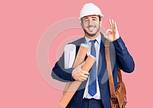 Young hispanic man wearing architect hardhat and leather bag holding blueprints doing ok sign with fingers, smiling friendly