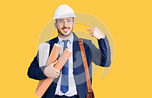Young hispanic man wearing architect hardhat and leather bag holding blueprints pointing finger to one self smiling happy and