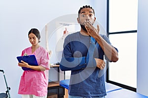 Young hispanic man wearing arm on sling at physiotherapy clinic covering mouth with hand, shocked and afraid for mistake