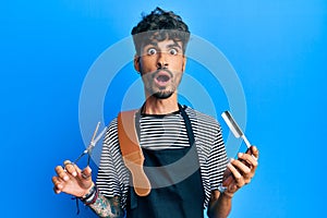 Young hispanic man wearing barber apron holding razor and scissors afraid and shocked with surprise and amazed expression, fear