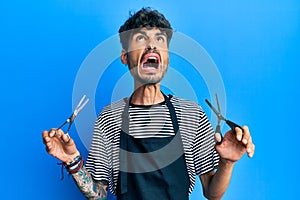 Young hispanic man wearing barber apron holding scissors angry and mad screaming frustrated and furious, shouting with anger