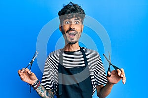 Young hispanic man wearing barber apron holding scissors celebrating crazy and amazed for success with open eyes screaming excited