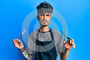 Young hispanic man wearing barber apron holding scissors skeptic and nervous, frowning upset because of problem