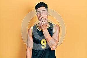 Young hispanic man wearing basketball uniform bored yawning tired covering mouth with hand