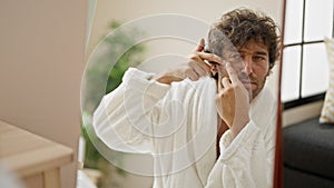 Young hispanic man wearing bathrobe popping pimple looking on mirror at home