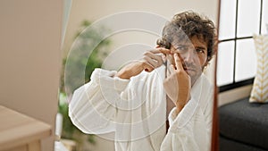 Young hispanic man wearing bathrobe popping pimple looking on mirror at home