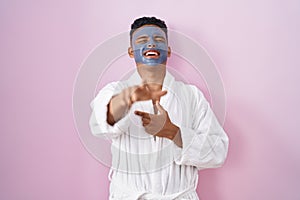 Young hispanic man wearing beauty face mask and bath robe laughing at you, pointing finger to the camera with hand over body,