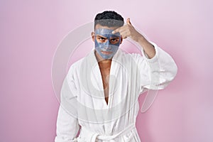 Young hispanic man wearing beauty face mask and bath robe pointing unhappy to pimple on forehead, ugly infection of blackhead