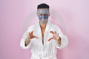 Young hispanic man wearing beauty face mask and bath robe smiling funny doing claw gesture as cat, aggressive and sexy expression